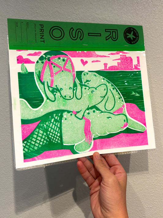 Hand holding a print with green and pink Manatee design on a gray surface